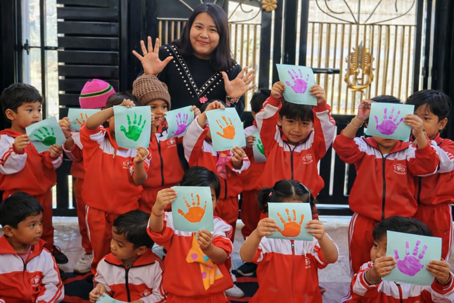 A group of cheerful young students in red and white tracksuits posing with their teacher. Each child is holding up a light blue piece of paper featuring a colorful handprint in shades of green, orange, or purple. The teacher stands in the center background with her hands raised, smiling at the camera. They are positioned outdoors in front of a black metal gate.