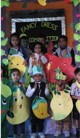 A group of young preschool-aged children participating in a "Fancy Dress Competition." The children are dressed in various fruit-themed costumes, including large paper cutouts of a red apple, yellow mangoes, and a green chili. One child in the center wears a black vest, a bow tie, and a paper crown while holding a star-tipped wand. They are standing in front of a black bulletin board decorated with paper leaves that reads "FANCY DRESS COMPETITION."
