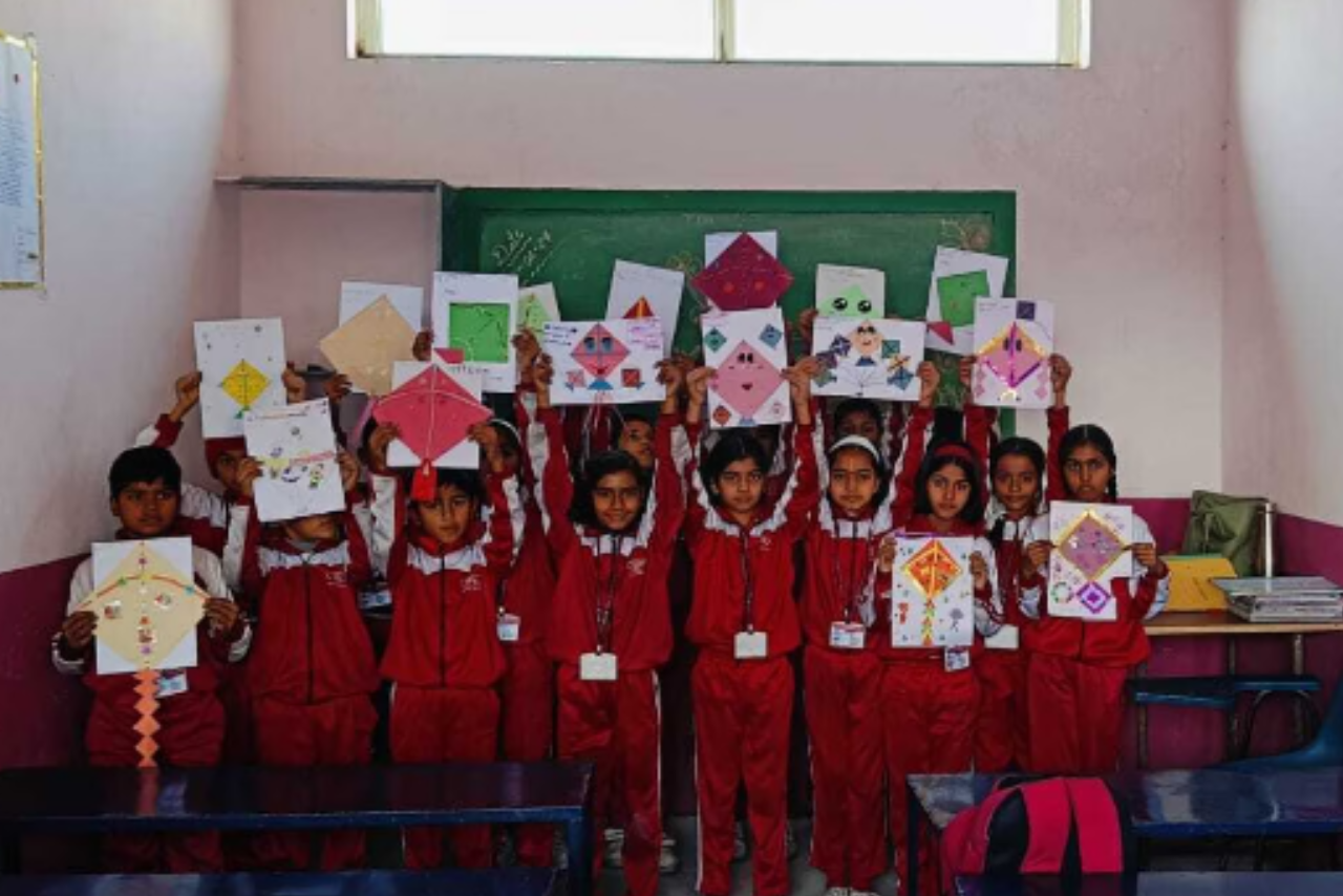A group of elementary-aged students in red and white tracksuits standing in a classroom, proudly holding up their handmade kite art. The kites are colorful and decorated with various patterns, tassels, and even small faces. They are posed in front of a green chalkboard, and the bright sunlight from a window above illuminates the room